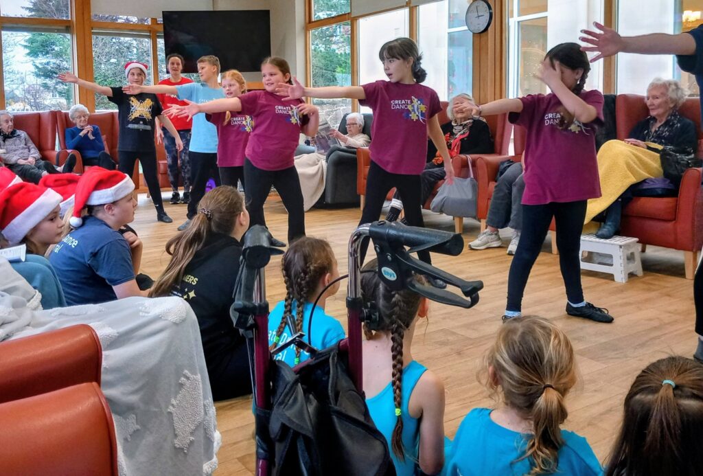 A group of children perform a dance for elderly residents seated in a care home, with some children dressed in costumes and others looking on.