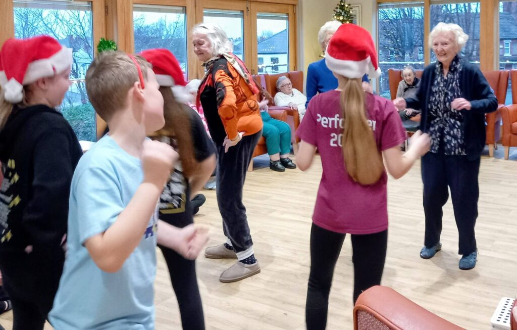 Children in Santa hats dance with elderly people in a brightly lit room, with a Christmas tree visible in the background.