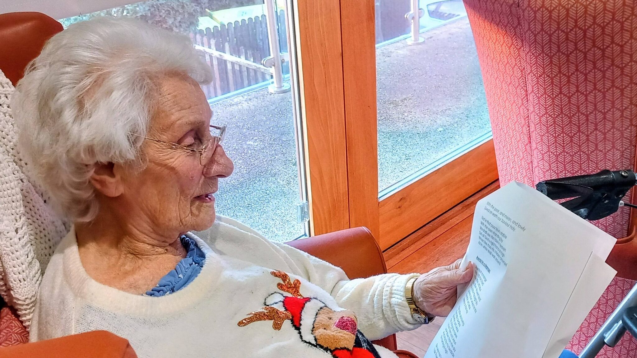 An elderly woman in a festive jumper sits in an armchair, reading printed sheets of paper by a window.