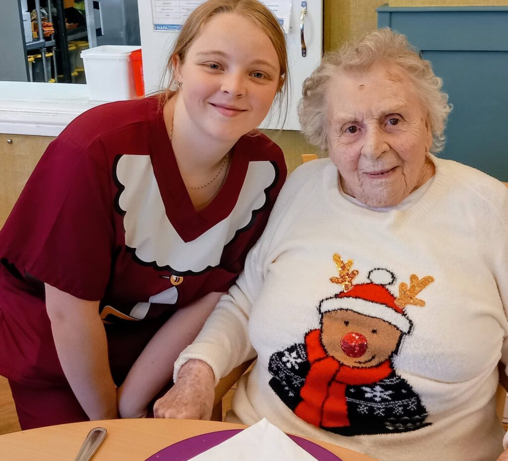 A young woman in a red festive top stands beside an older woman wearing a white jumper with a reindeer design. Both are smiling at the camera.