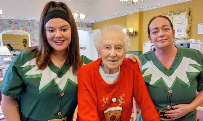 An elderly woman in a red reindeer jumper stands between two women in green elf costumes, all smiling at the camera in a festively decorated indoor setting.