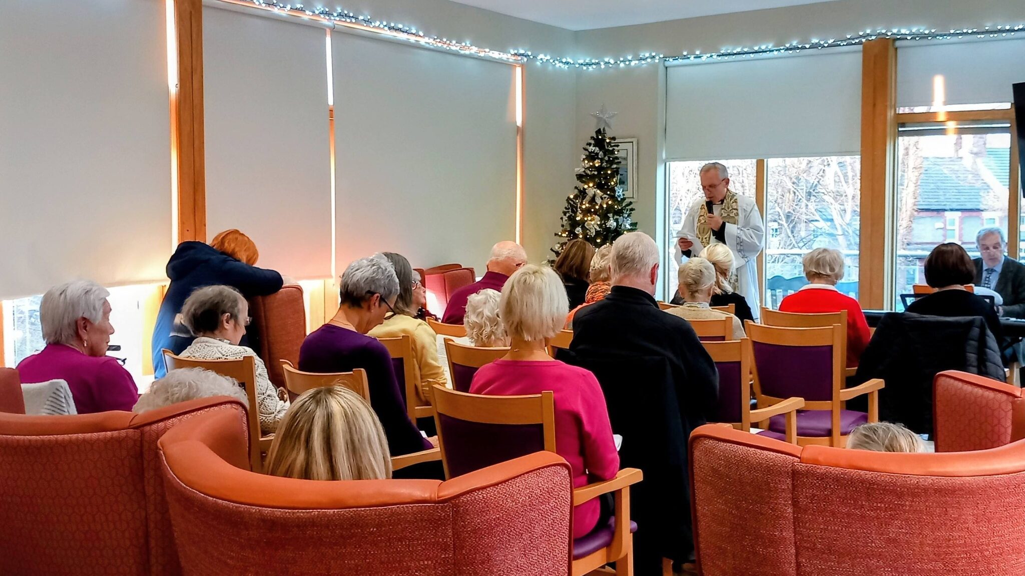 A group of elderly people are seated in a room, facing a man who is speaking at the front near a decorated Christmas tree.