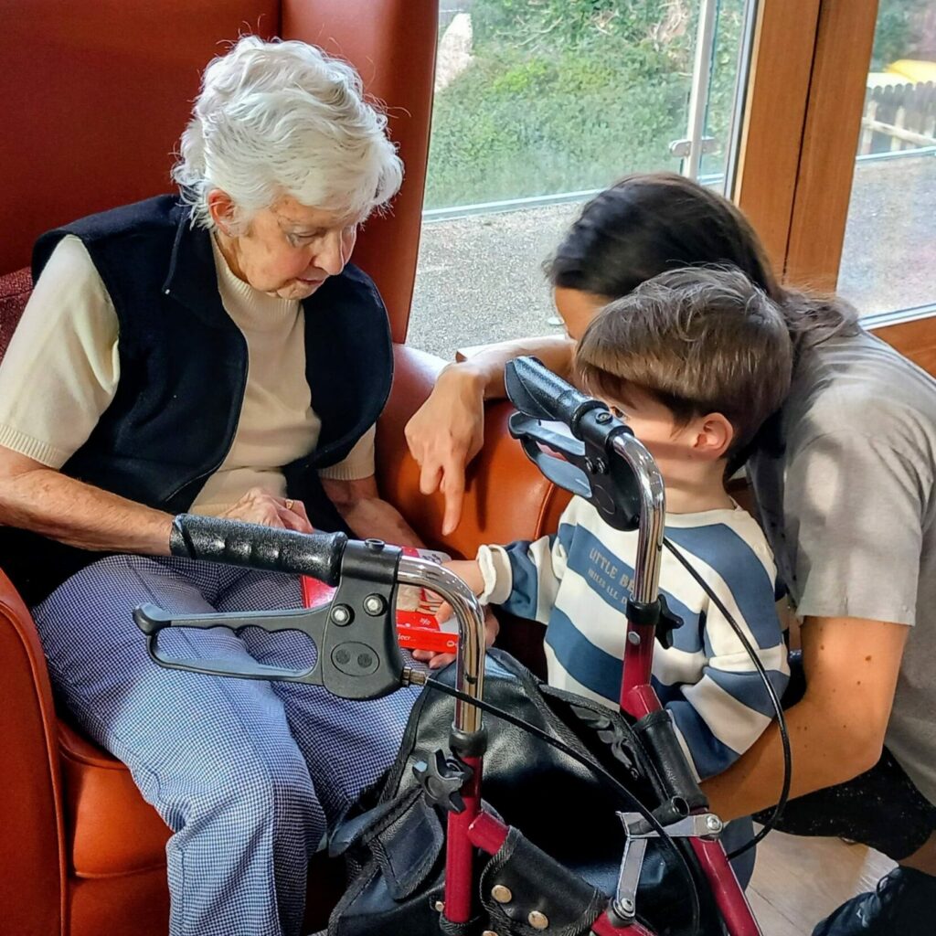 An elderly woman sits in an armchair, engaging with a young boy and a woman beside her; a walking frame rests in front of her knees as soft Christmas music plays in the background.