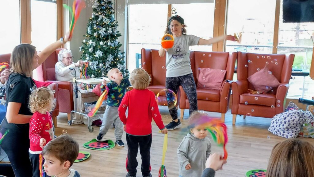 A woman leads a group of children in a ribbon dance activity indoors with music playing, bubbles floating about, and a decorated Christmas tree in the background while some adults and Popcats sit and watch.