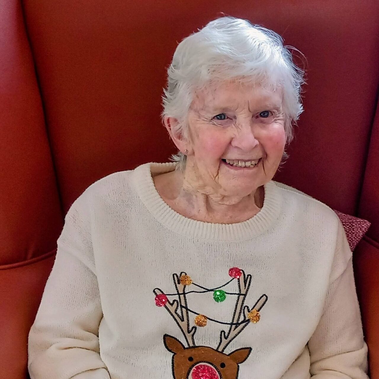 Elderly woman with white hair sits on a red chair, smiling, wearing a cream jumper with a reindeer and Christmas lights design, and checked trousers.