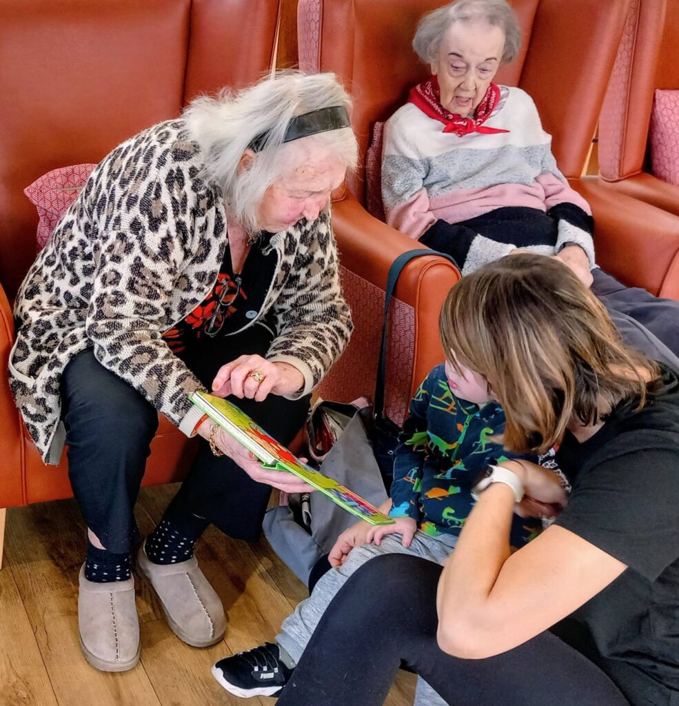 An elderly woman shows a book to a child while another elderly woman and an adult sit nearby on orange chairs, with Christmas music gently playing in the background.