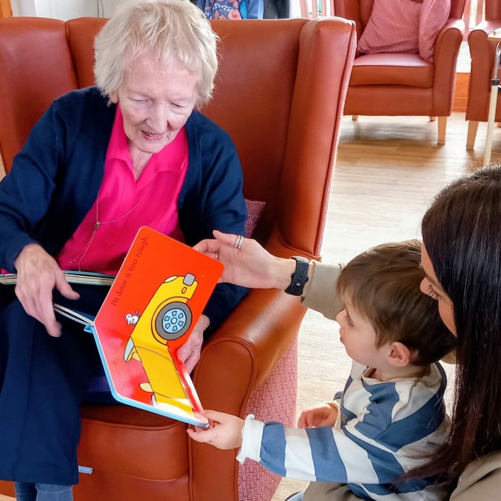 An elderly woman sits in an armchair, reading a colourful Christmas picture book to a young child, as another adult helps the child hold the book.