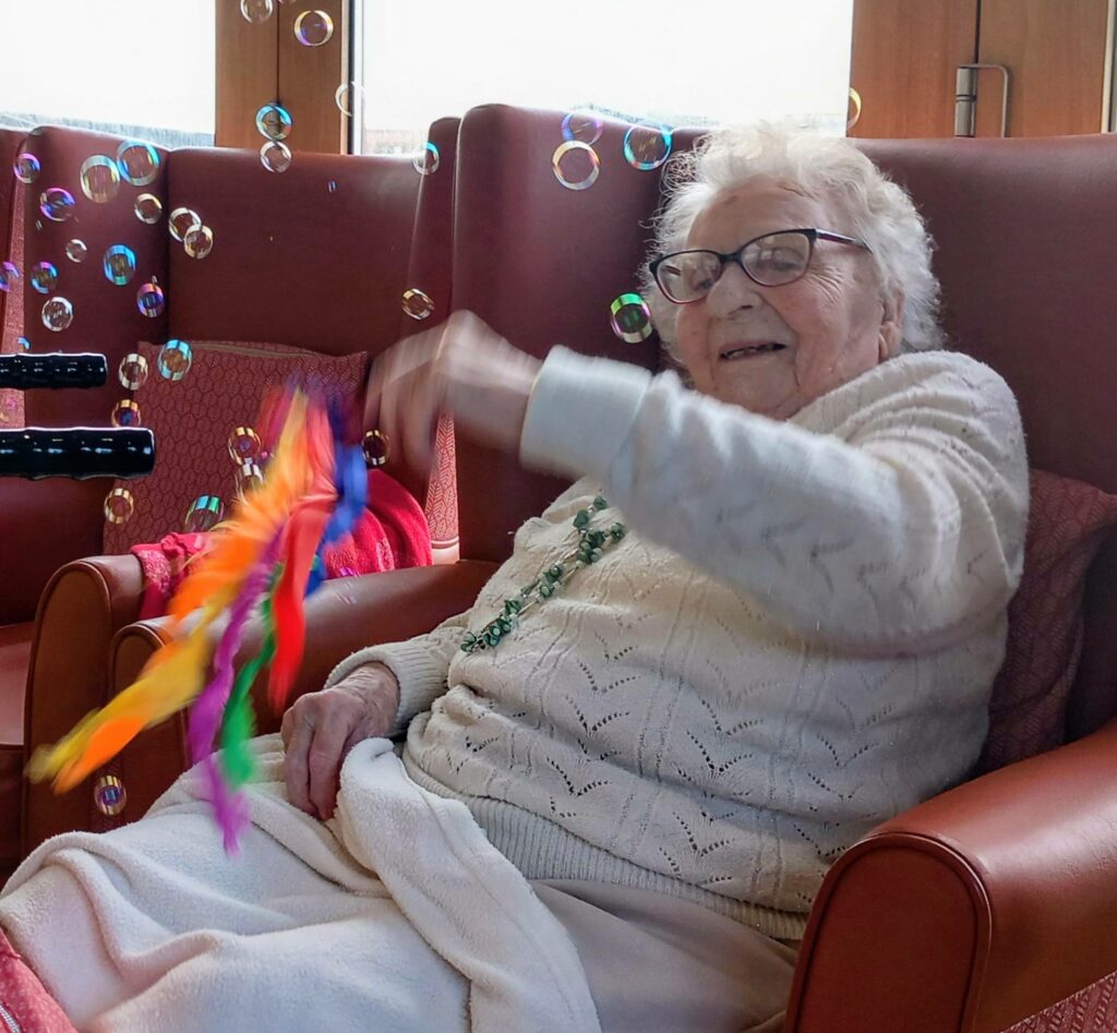 An elderly woman sits in an armchair, waving a colourful scarf and smiling as bubbles float about her in a bright room, with cheerful Christmas music playing softly in the background.