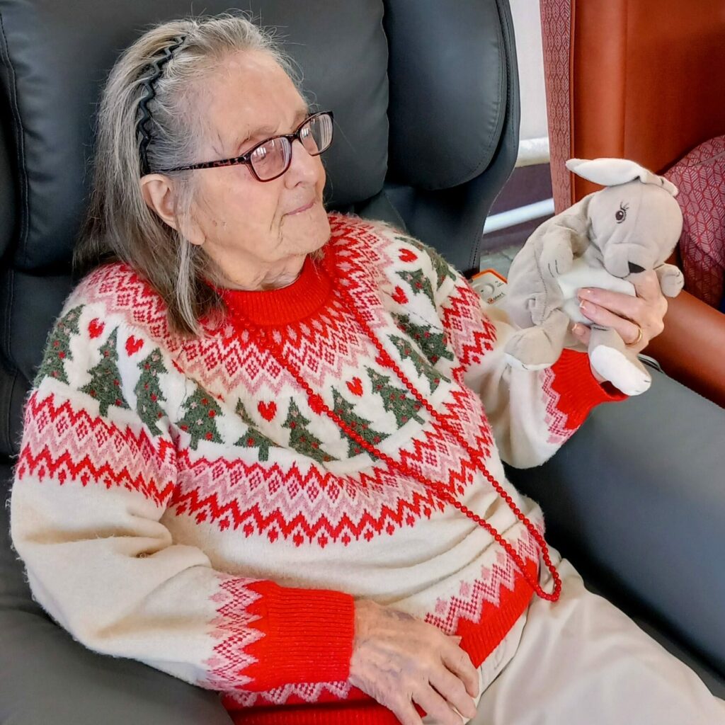 Elderly woman wearing spectacles and a festive jumper sits in an armchair, holding a small cuddly toy in her left hand, as Christmas Popcats music brings a smile to her face.
