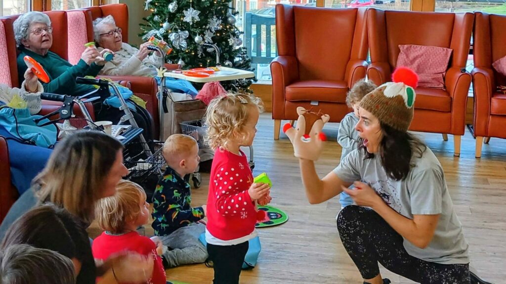 A woman in festive clothing entertains a group of smiling children and elderly people in a room with a Christmas tree, large windows, and cheerful music playing on a bright morning.