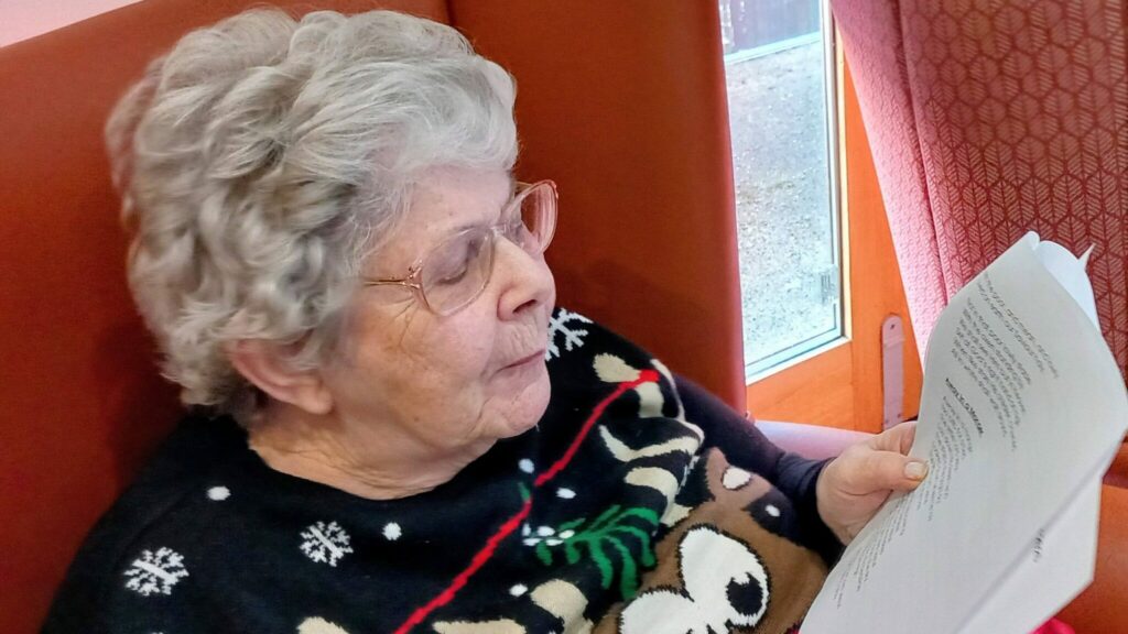 Elderly woman with white hair and glasses sits in a chair, wearing a reindeer jumper, and reads from a stack of printed papers.
