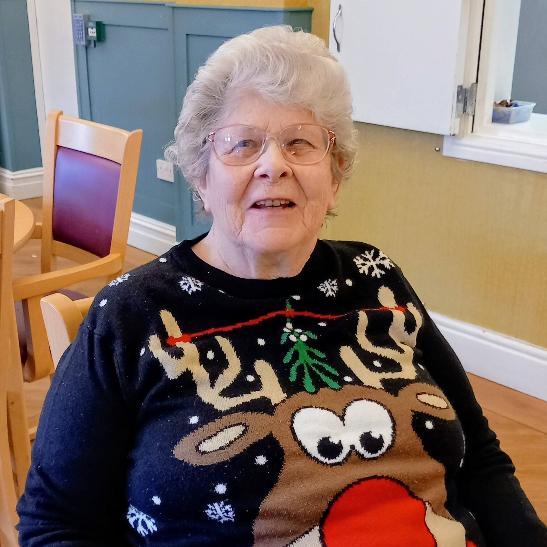 An elderly woman with spectacles is sitting indoors and smiling, wearing a black Christmas jumper with a reindeer design.