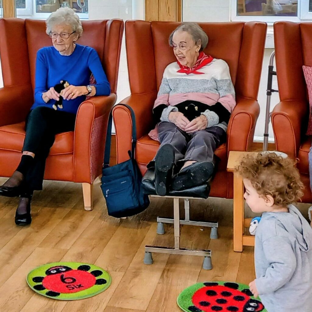Two elderly women sit in armchairs holding cuddly toys, while a small child stands nearby on a floor decorated with colourful ladybird mats, creating a heartwarming scene reminiscent of Christmas.
