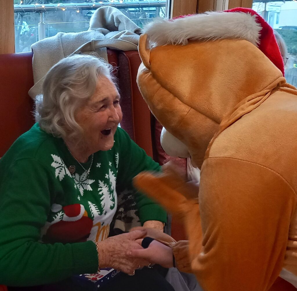 An elderly woman in a festive jumper smiles and holds hands with someone in a reindeer costume inside a warmly lit room.