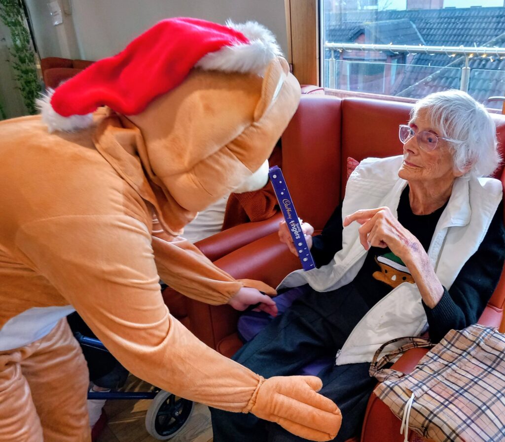 A person in a reindeer costume hands a chocolate bar to an elderly woman sitting on a red sofa.
