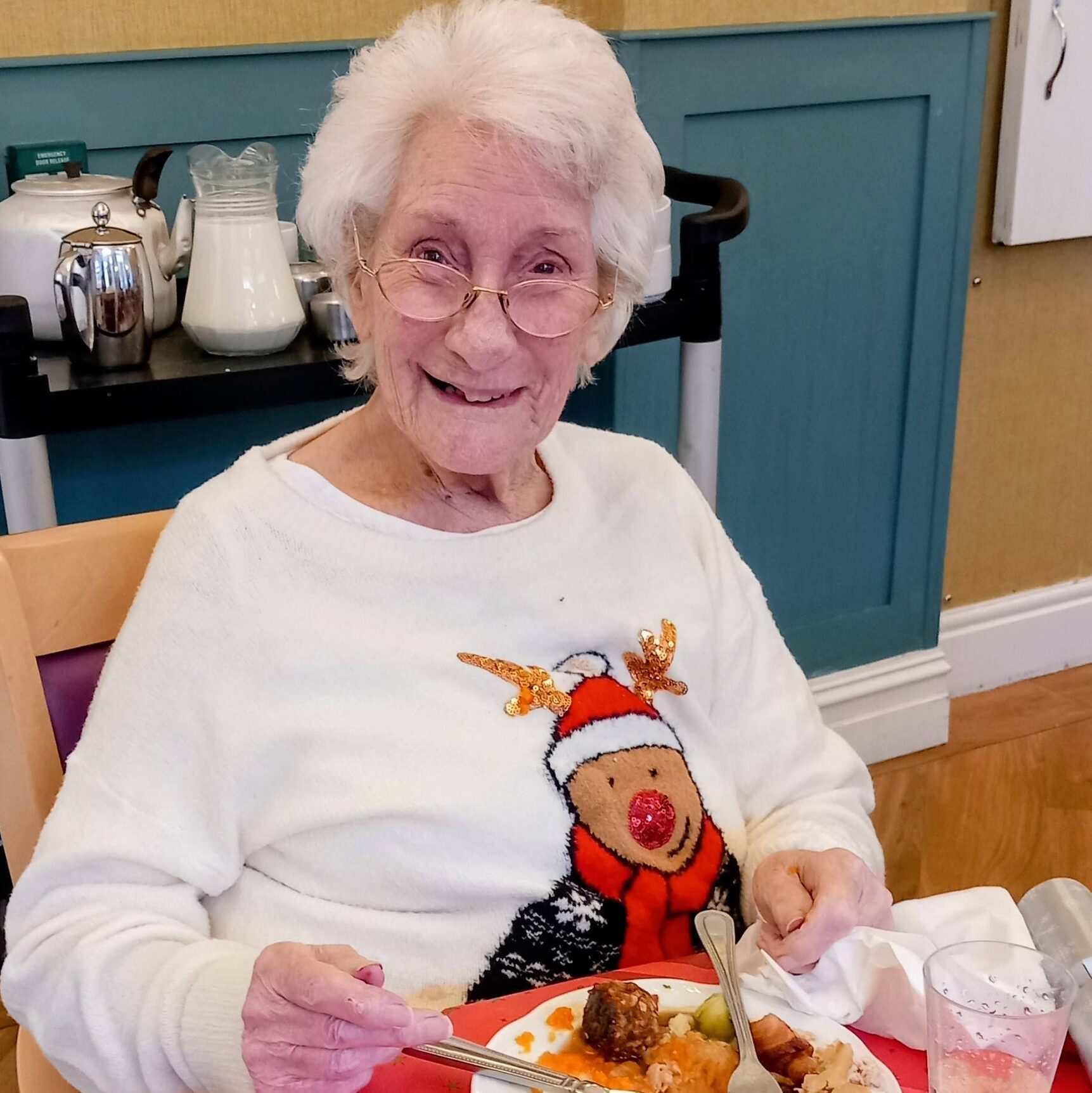 Elderly woman with white hair, wearing a festive reindeer jumper, smiling as she eats a meal at a table set with orange juice and a red tablecloth.