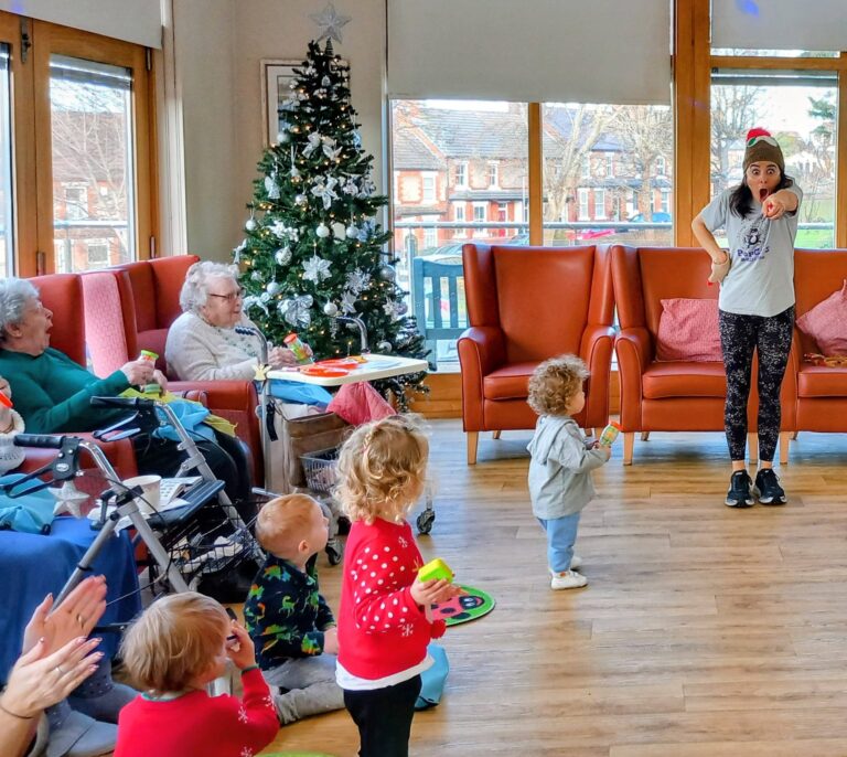 A group of children and elderly people enjoy a music session beside a decorated Christmas tree, with a woman leading the festive activity.