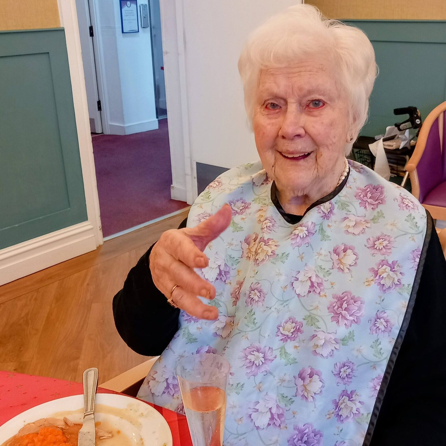 An elderly woman wearing a floral bib sits at a table with an empty plate and a glass, gesturing with one hand and smiling at the camera.