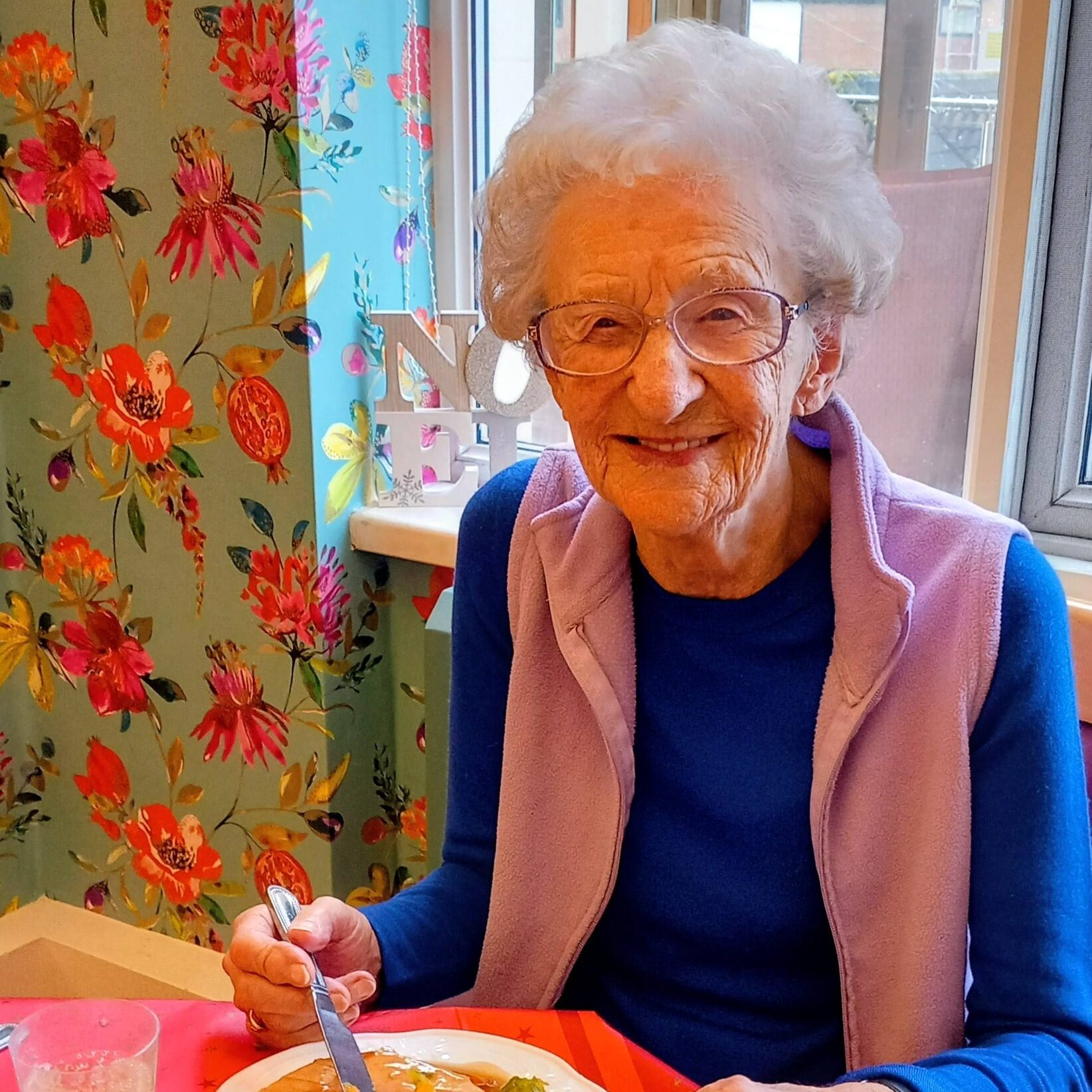 Elderly woman with spectacles and white hair smiles while enjoying a meal at a table in a brightly decorated room with floral wallpaper.