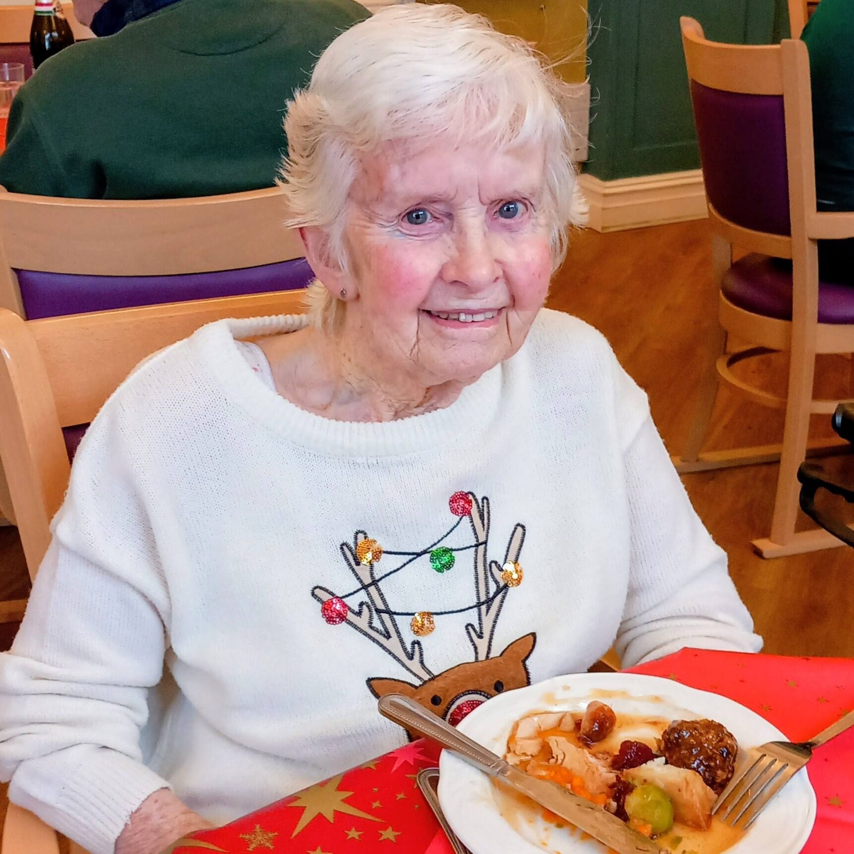 An elderly woman with white hair sits at a table covered with a festive red tablecloth, wearing a reindeer jumper and eating a meal.