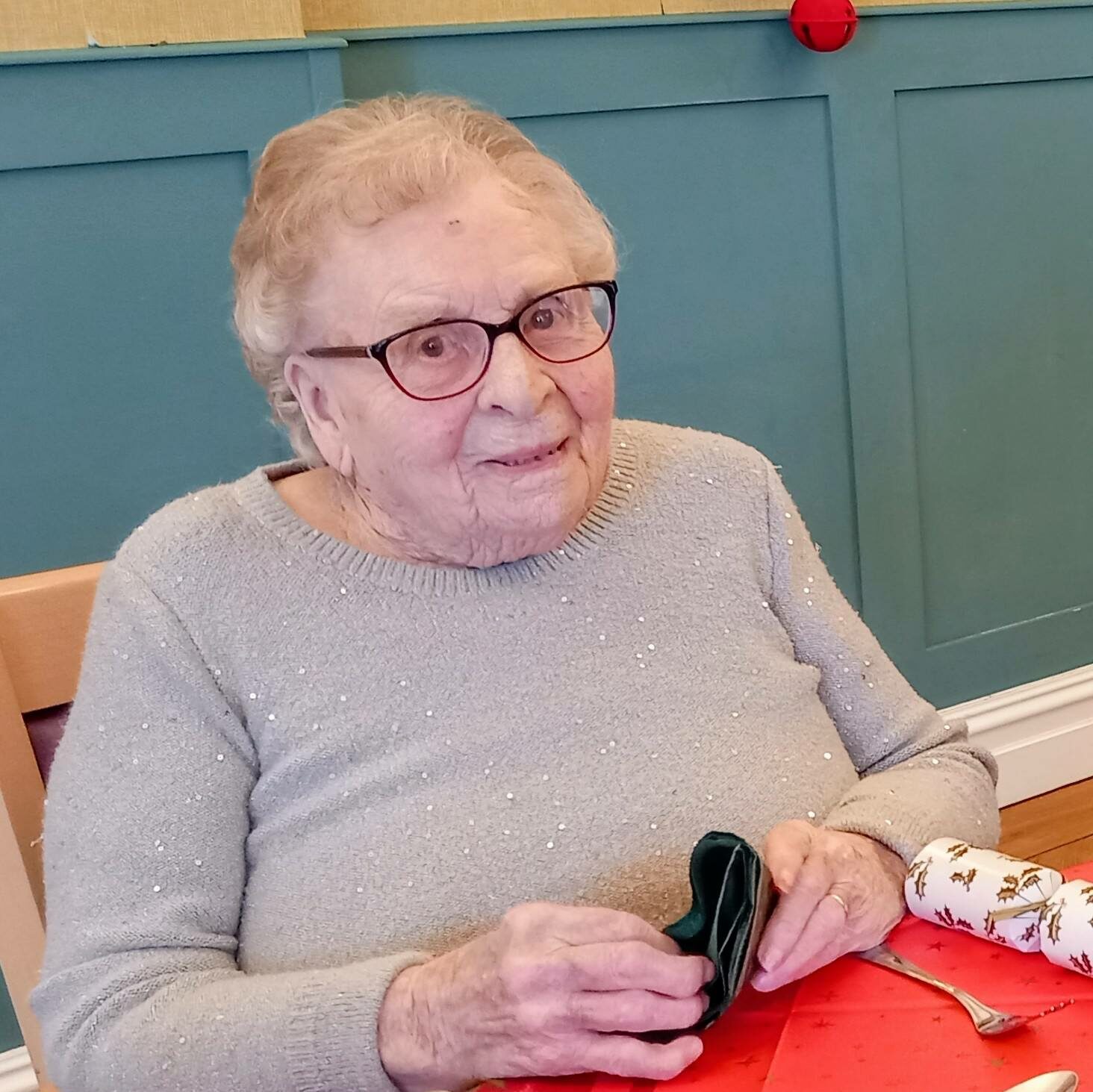An elderly woman wearing glasses sits at a table laid for a meal, holding a green serviette and looking towards the camera.
