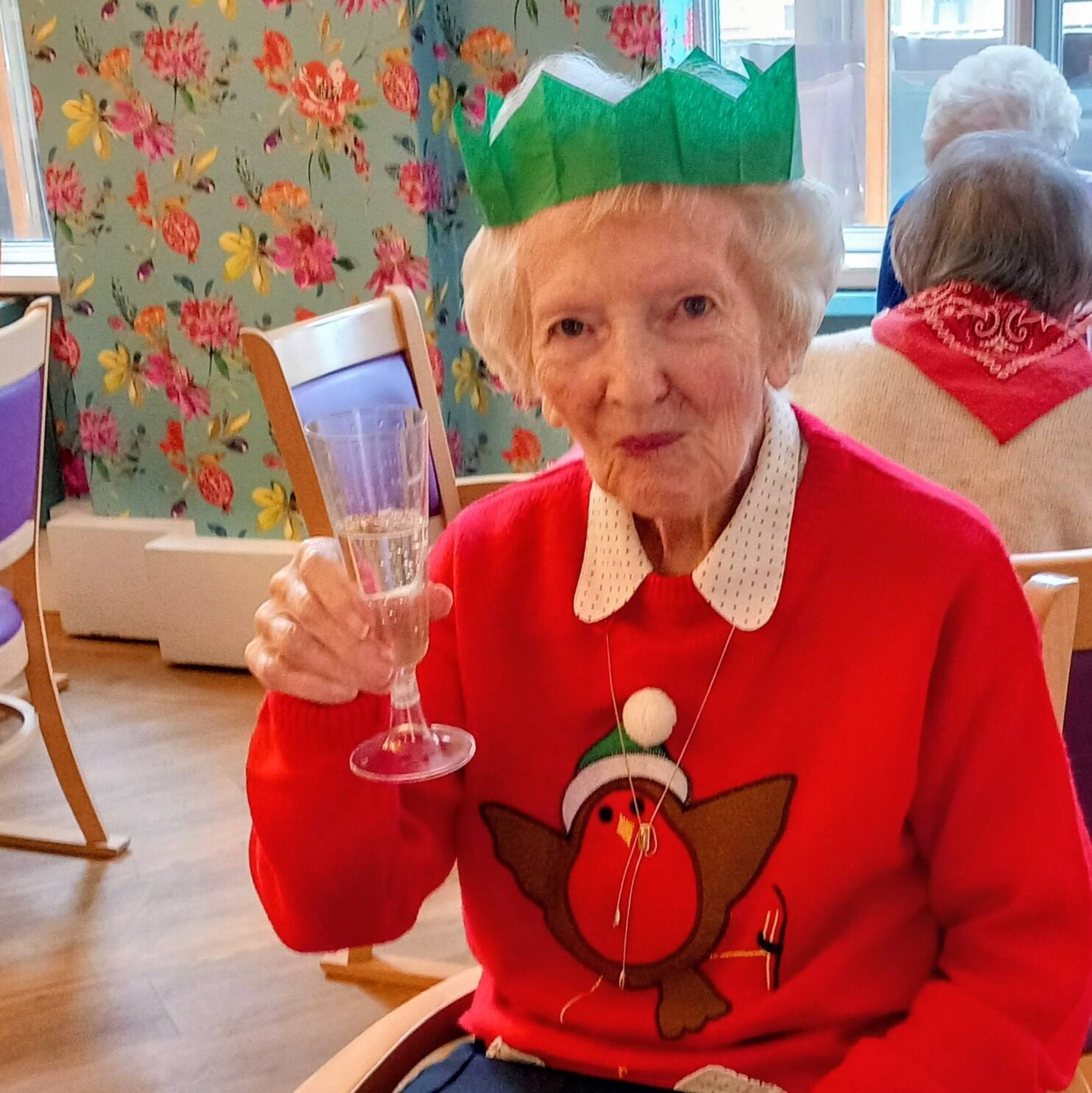 An elderly woman in a festive jumper and green paper crown raises a glass at a table laid with a Christmas meal.