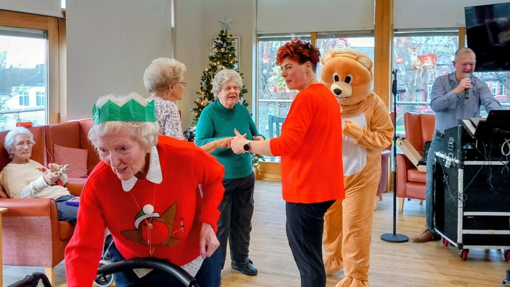 A group of elderly people and someone in a bear costume enjoy a festive gathering in a decorated room, featuring a Christmas tree and a man singing by the sound system.