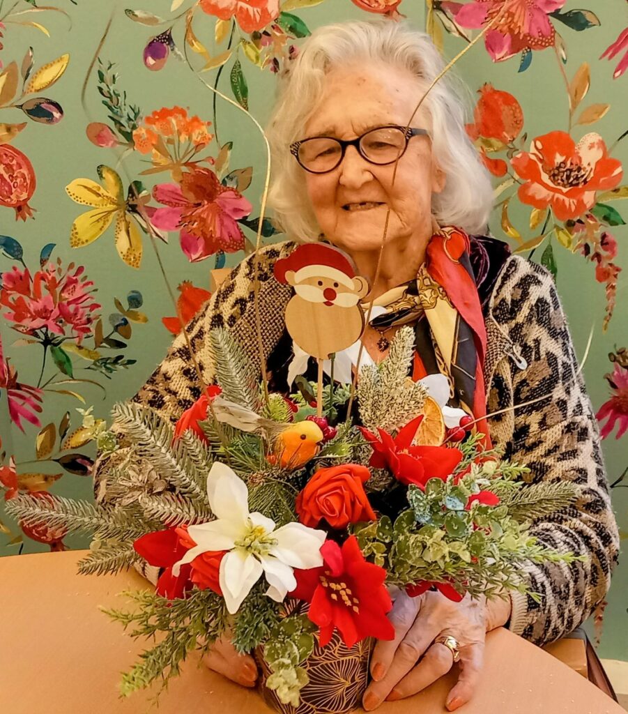An elderly woman with white hair and spectacles sits at a table with a festive floral arrangement in front of her, set against a colourful floral wallpaper backdrop.