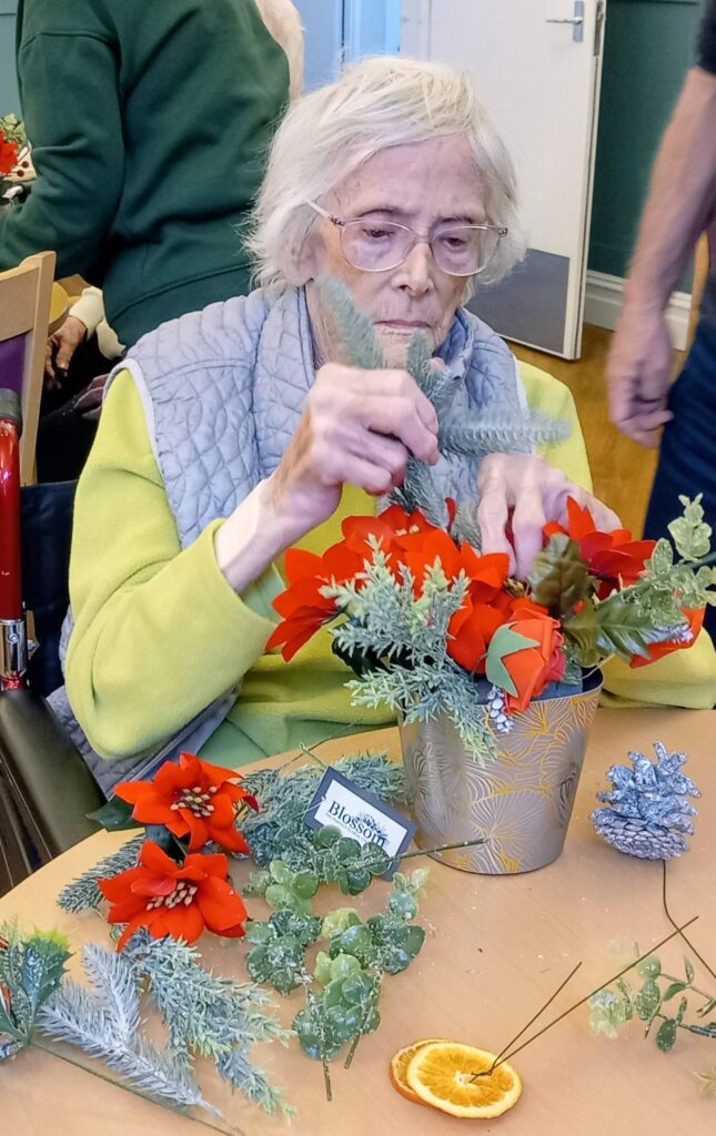 An elderly woman arranges artificial red flowers and greenery in a gold pot on a table, with craft materials scattered nearby.