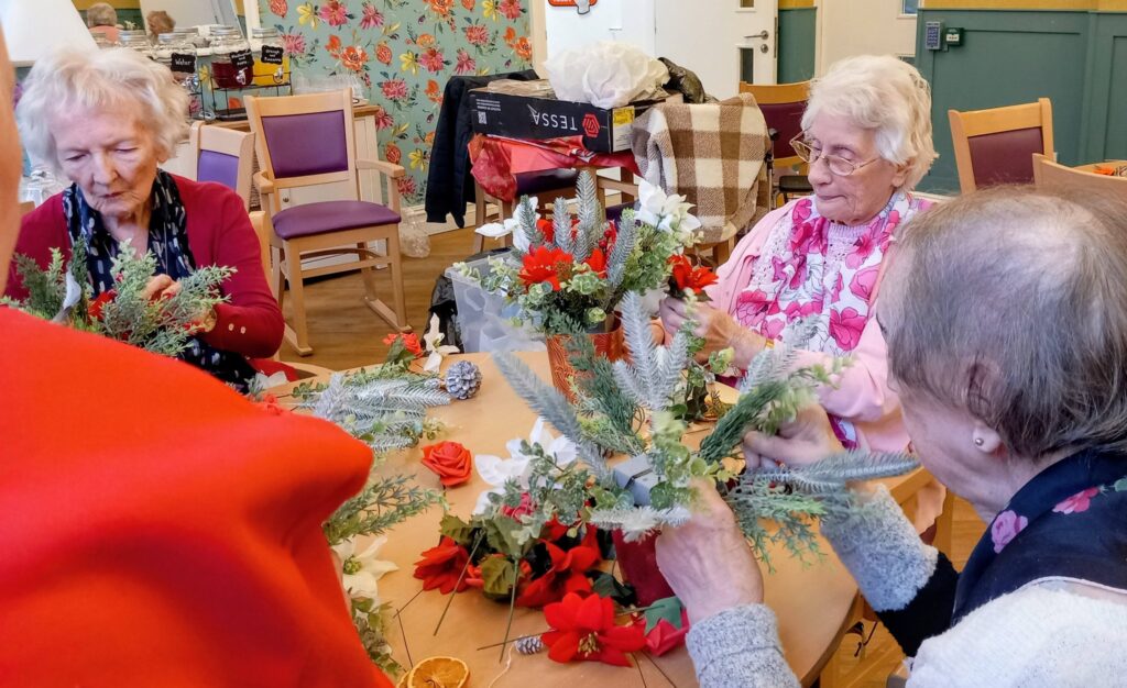Three elderly women are seated at a table making floral arrangements with red, white, and green decorations in a brightly lit room.