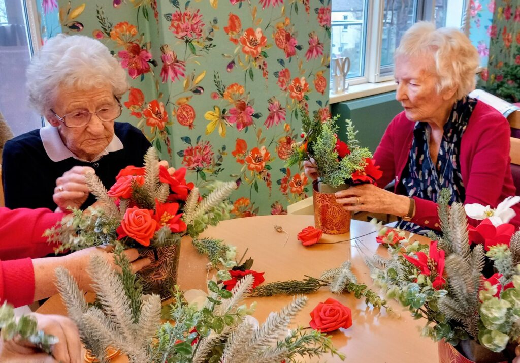 Two elderly women sit at a table arranging red flowers and greenery in vases, with bright floral wallpaper and a window in the background.