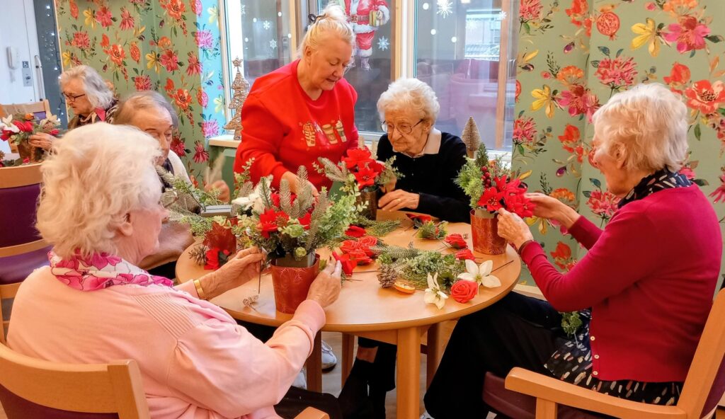 A group of elderly women sit around a table creating festive floral arrangements in a brightly decorated room.