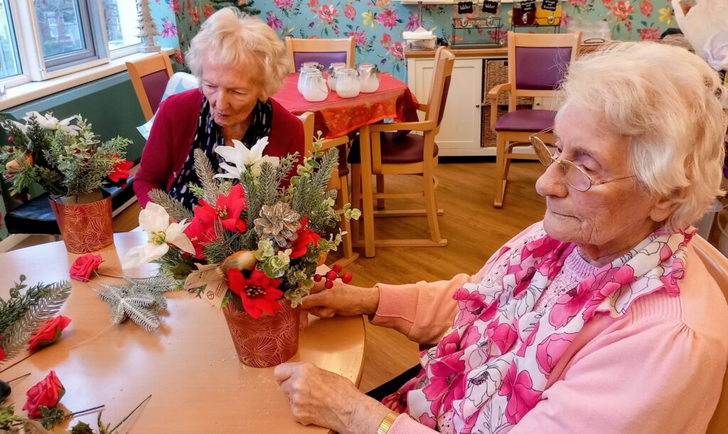 Two elderly women seated at a table in a brightly decorated room, arranging artificial flowers in vases.