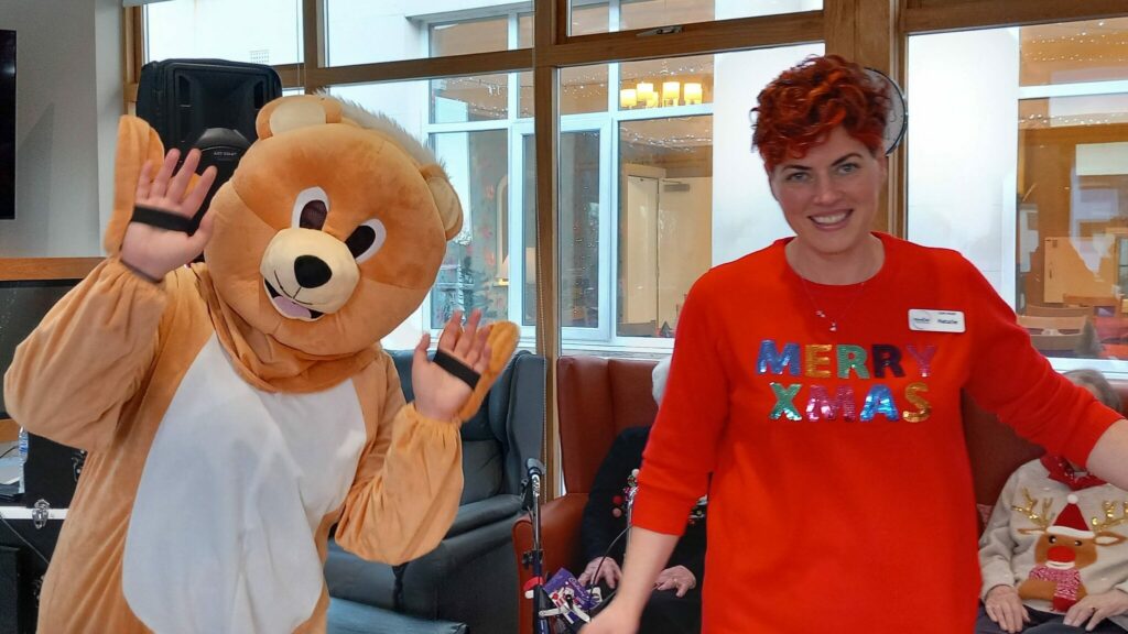 A person in a bear costume stands beside a woman wearing a red Merry Xmas jumper in a room with people seated in the background.