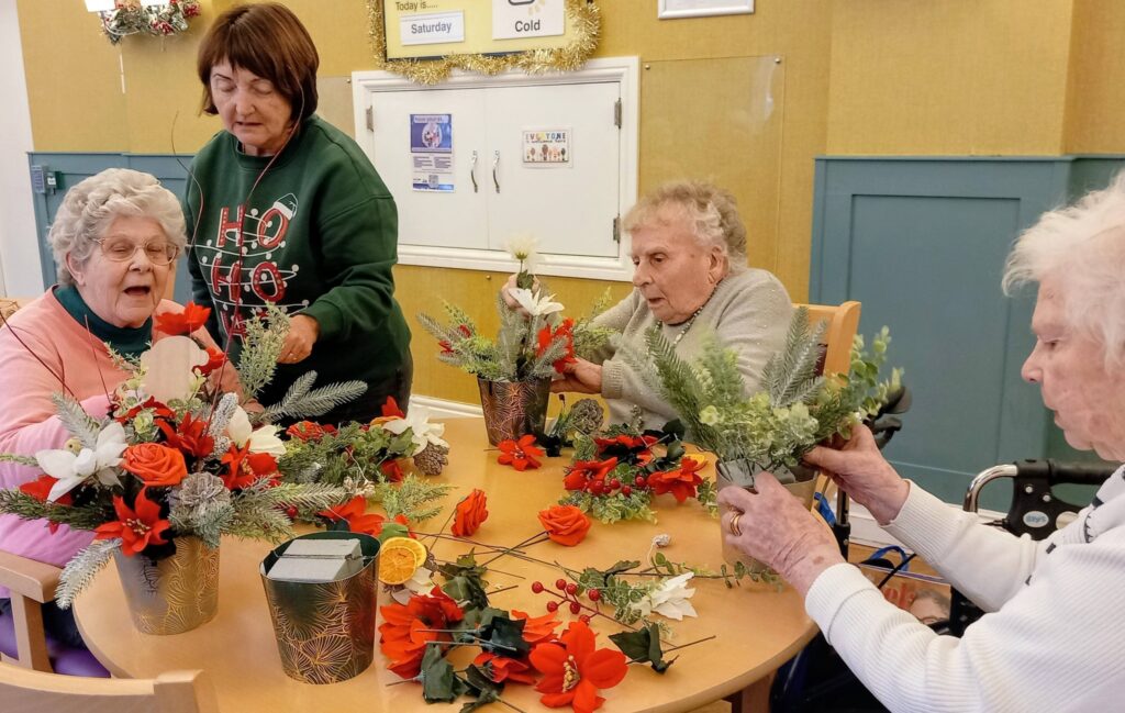 Four elderly women sit around a table arranging flowers and greenery in vases in a brightly lit communal room decorated for the festive season.