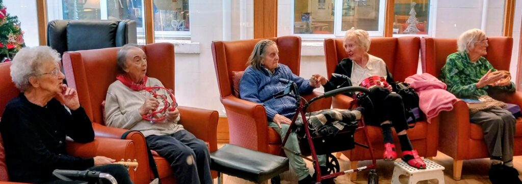 Five elderly women sit in armchairs in a communal lounge, some holding musical instruments and blankets, enjoying music and conversation as part of the enjoyable activities.