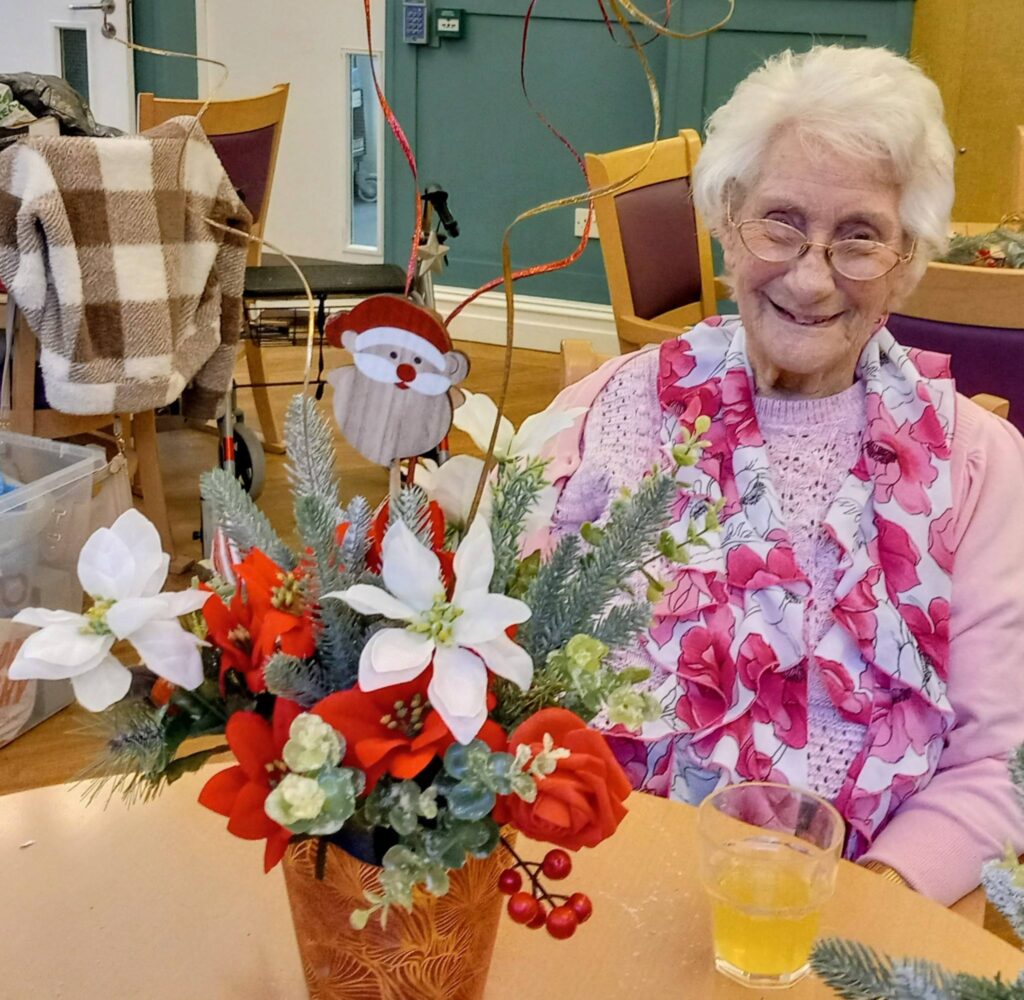 An elderly woman with white hair and glasses sits at a table with a festive floral arrangement and a glass of orange juice in front of her.