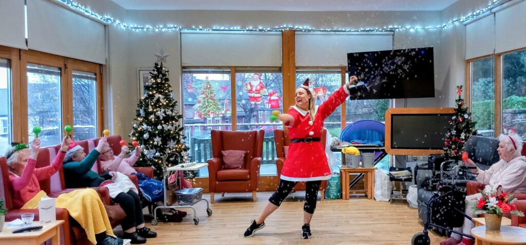 A woman dressed as Santa leads a lively exercise session with music for a group of elderly people in a decorated room adorned with Christmas trees and fairy lights.