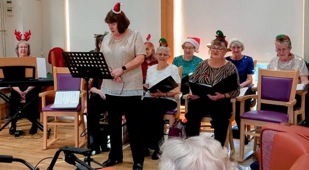 A group of elderly women, some wearing Christmas hats and antlers, enjoy music as they sit and sing in a brightly lit room, with one woman standing and reading from a music stand.