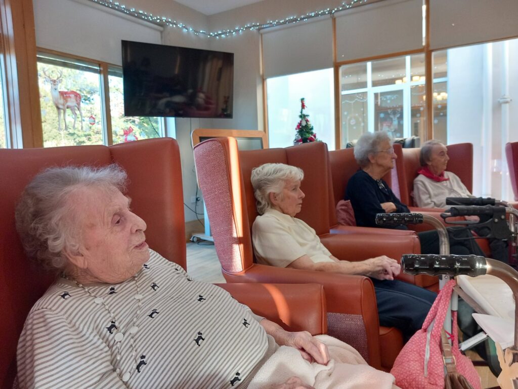 Four elderly women sit in cushioned chairs in a brightly lit communal room, soft music playing, with walking frames close by and a Christmas tree visible in the background.