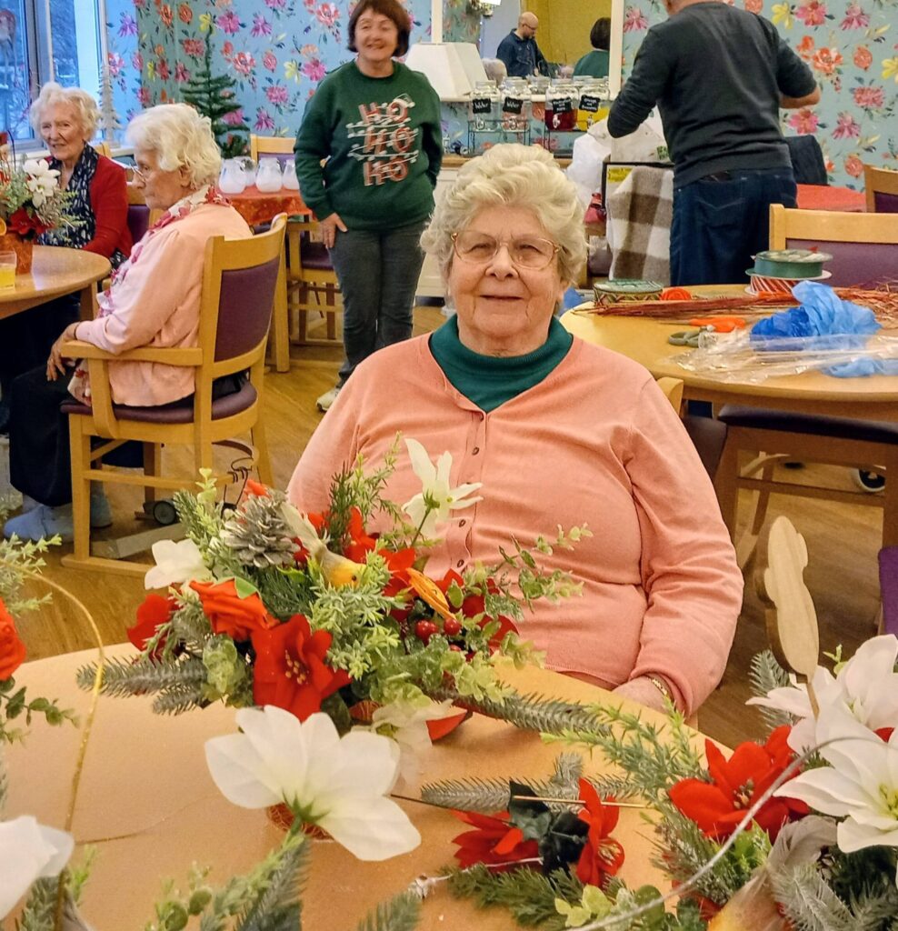 An elderly woman sits at a table adorned with festive flower arrangements in a community room, as others socialise and work in the background.