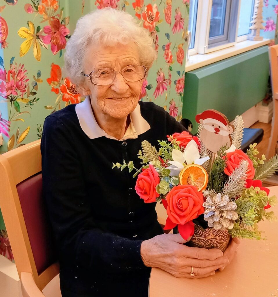An elderly woman wearing glasses sits at a table, holding a festive floral arrangement featuring red flowers, pinecones, and a Father Christmas decoration.