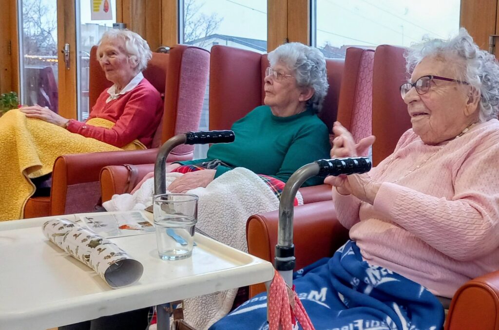 Three elderly women sit in armchairs in a bright room, covered with blankets, as soft music plays. A walking frame, a glass of water, and a rolled-up magazine are on a nearby table.