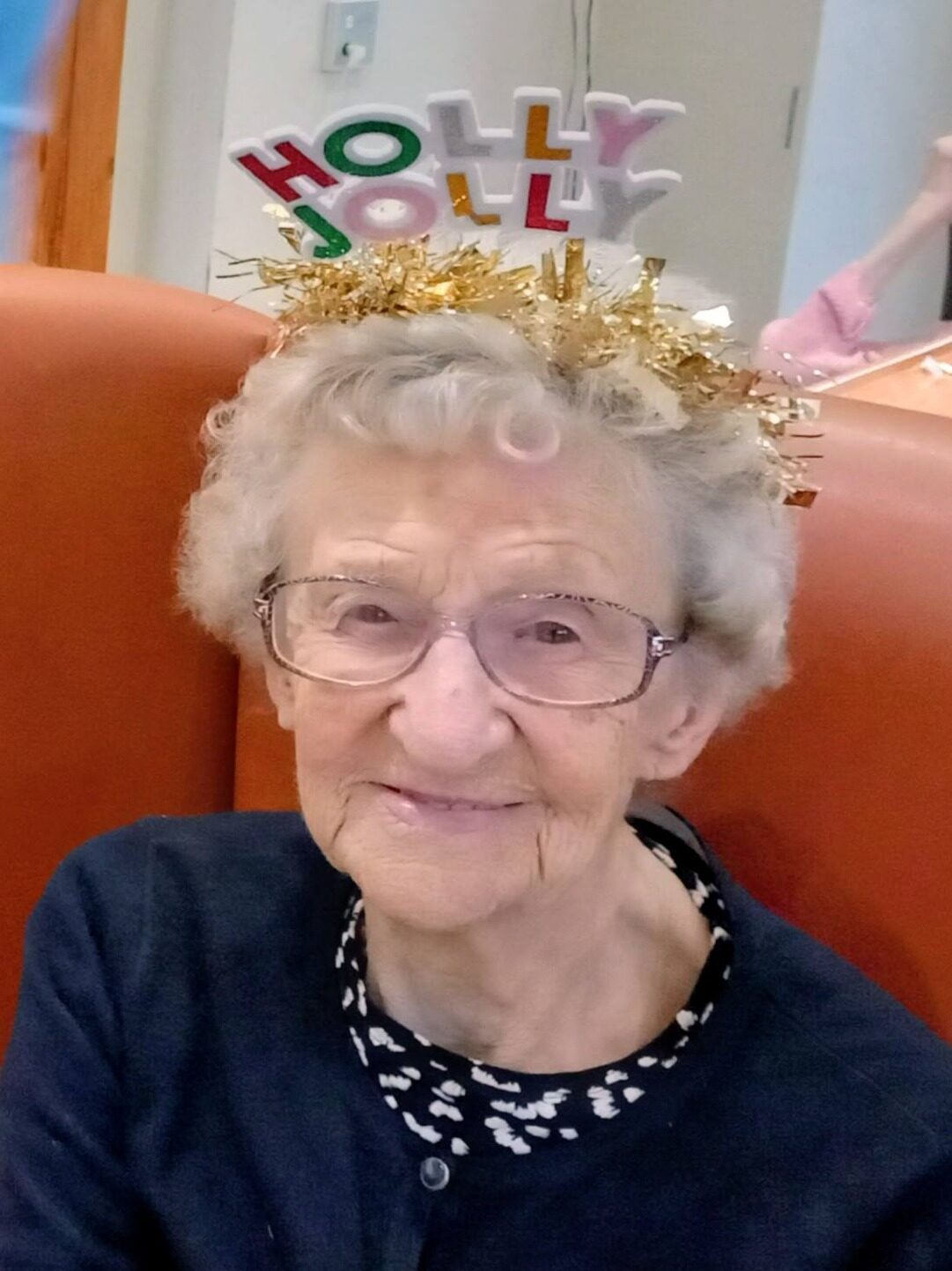Elderly woman with spectacles and curly grey hair smiles while wearing a festive Holly Jolly headband and a dark cardigan, ready to enjoy fun activities and music.