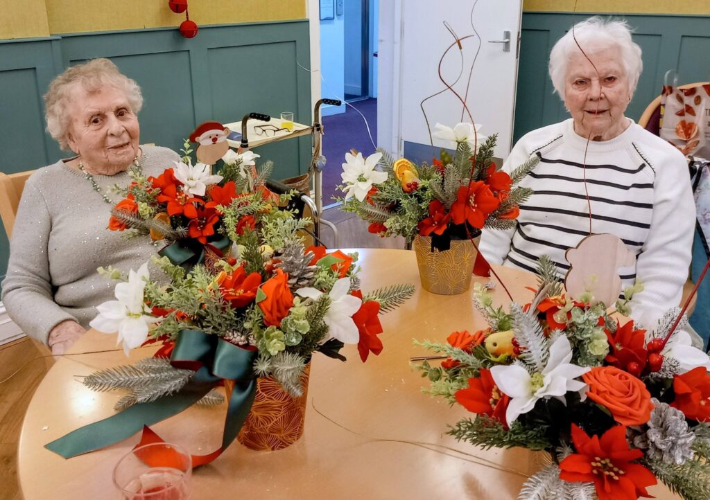 Two elderly women sit at a table with festive floral arrangements featuring red and white flowers, greenery, and decorative elements in a communal indoor setting.