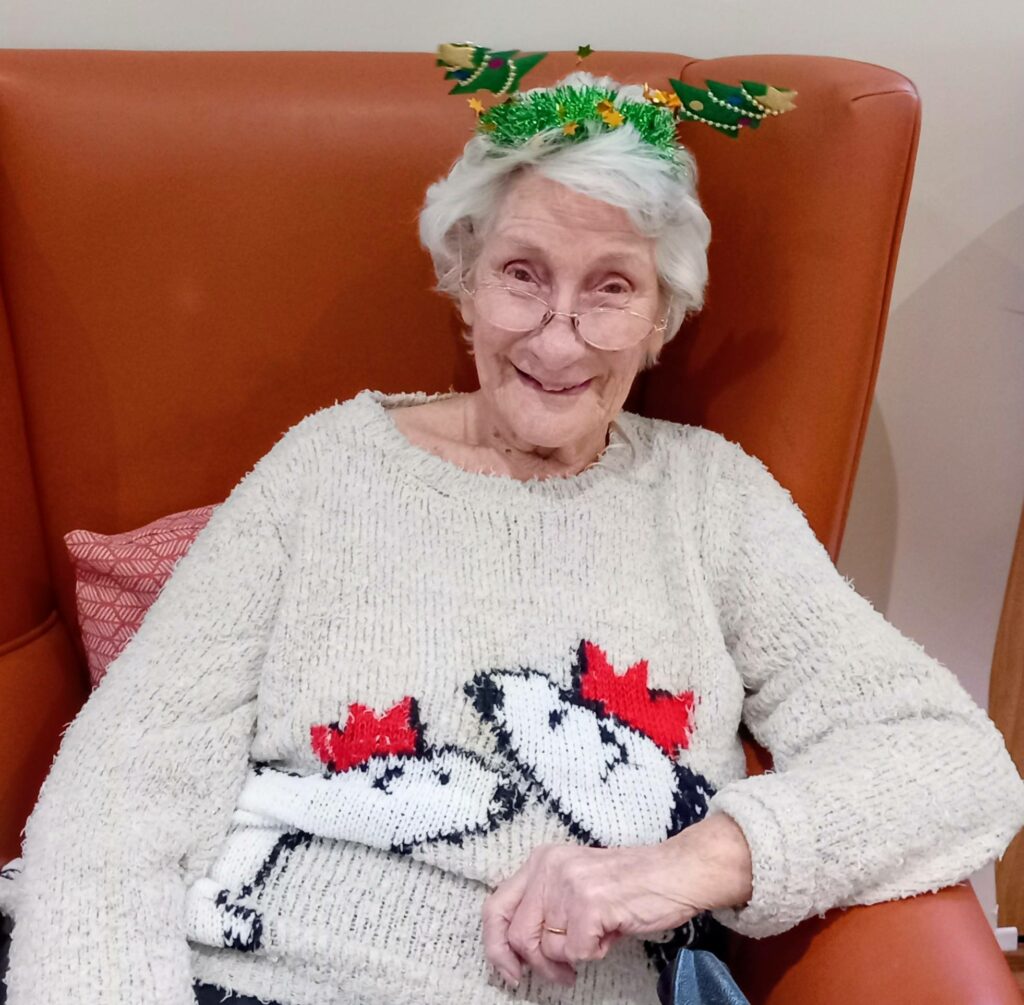 An elderly woman with white hair, wearing glasses, a festive headband, and a Snoopy jumper with red hats, sits in an orange chair and smiles, looking ready for fun activities this holiday season.