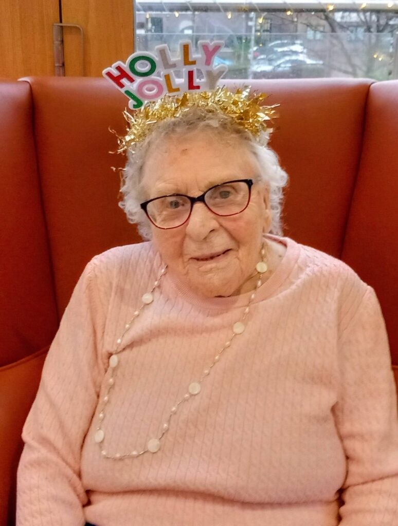 Elderly woman wearing glasses, a pink jumper, and a necklace sits on an orange chair, sporting a gold tinsel 'Holly Jolly' headband—ready for fun activities and festive run music.