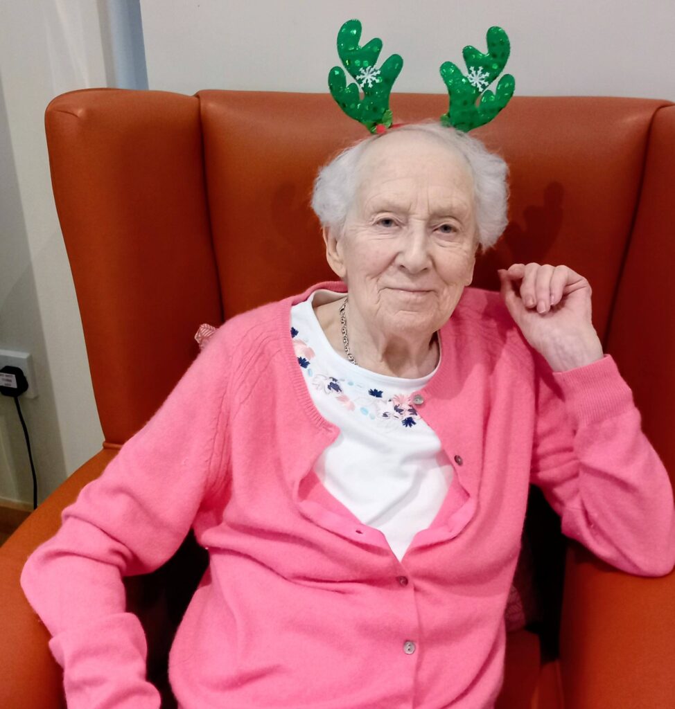 Elderly woman seated in an orange armchair, dressed in a pink cardigan and green reindeer antler headband, smiling at the camera during festive musical activities.