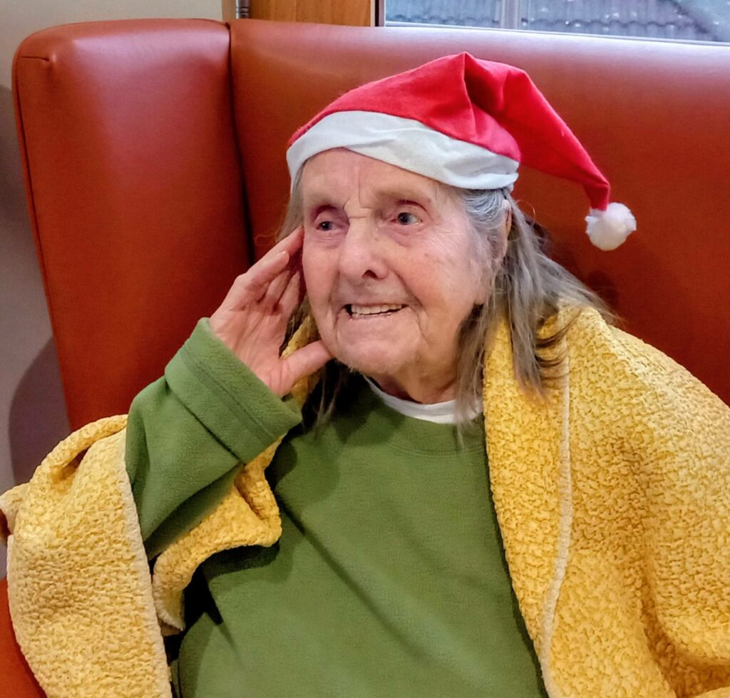Elderly woman wearing a red Father Christmas hat, green top, and yellow blanket, seated on an orange chair and smiling as she enjoys festive music.
