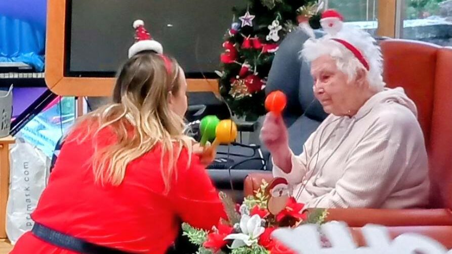A woman in red and an elderly lady, both wearing festive headbands, shake maracas near a decorated Christmas tree, enjoying music and fun activities in a cosy room.